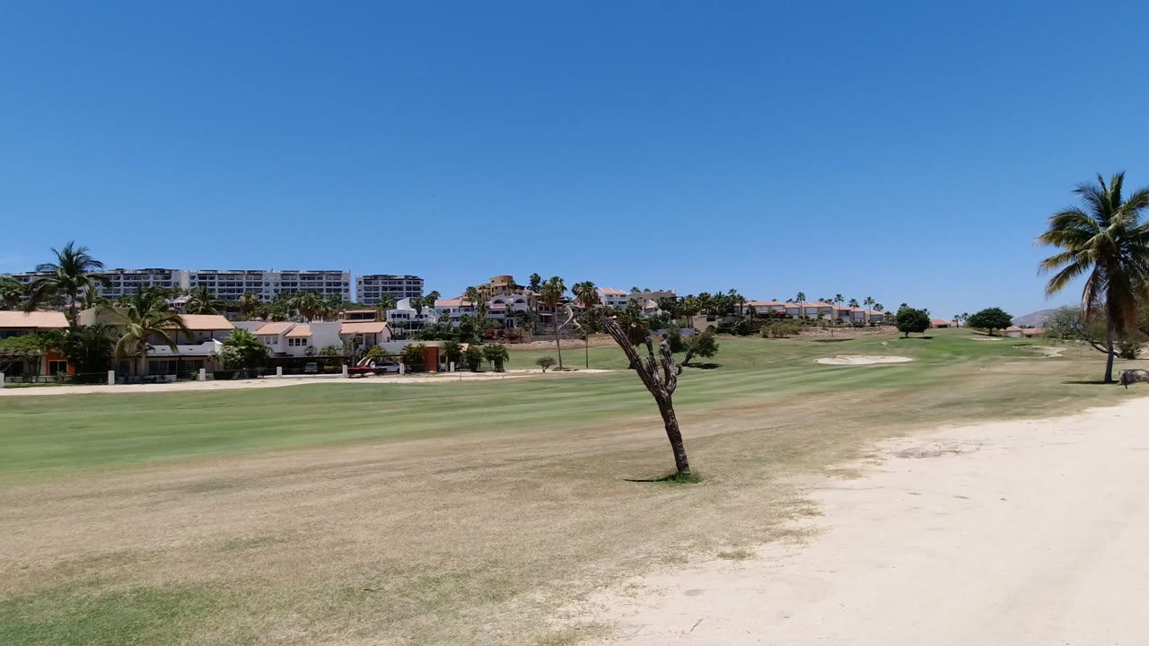Wide view of a Los Cabos golf course with sandy ground and fairway grass in front, palms and low homes beyond, distant buildings on the horizon, and no visible people.