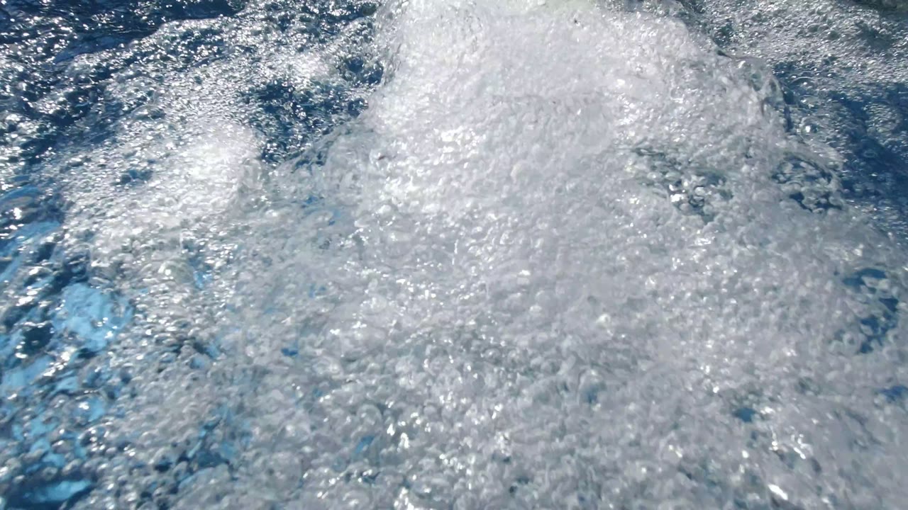 Foamy white bubbles spread over aqua hot tub water in Los Cabos, Mexico, with bright surface reflections and no visible people.