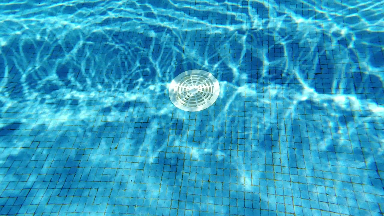 Overhead view of turquoise pool water in Los Cabos, Mexico, with bright sun reflections over blue tile and a white circular drain, no visible people.