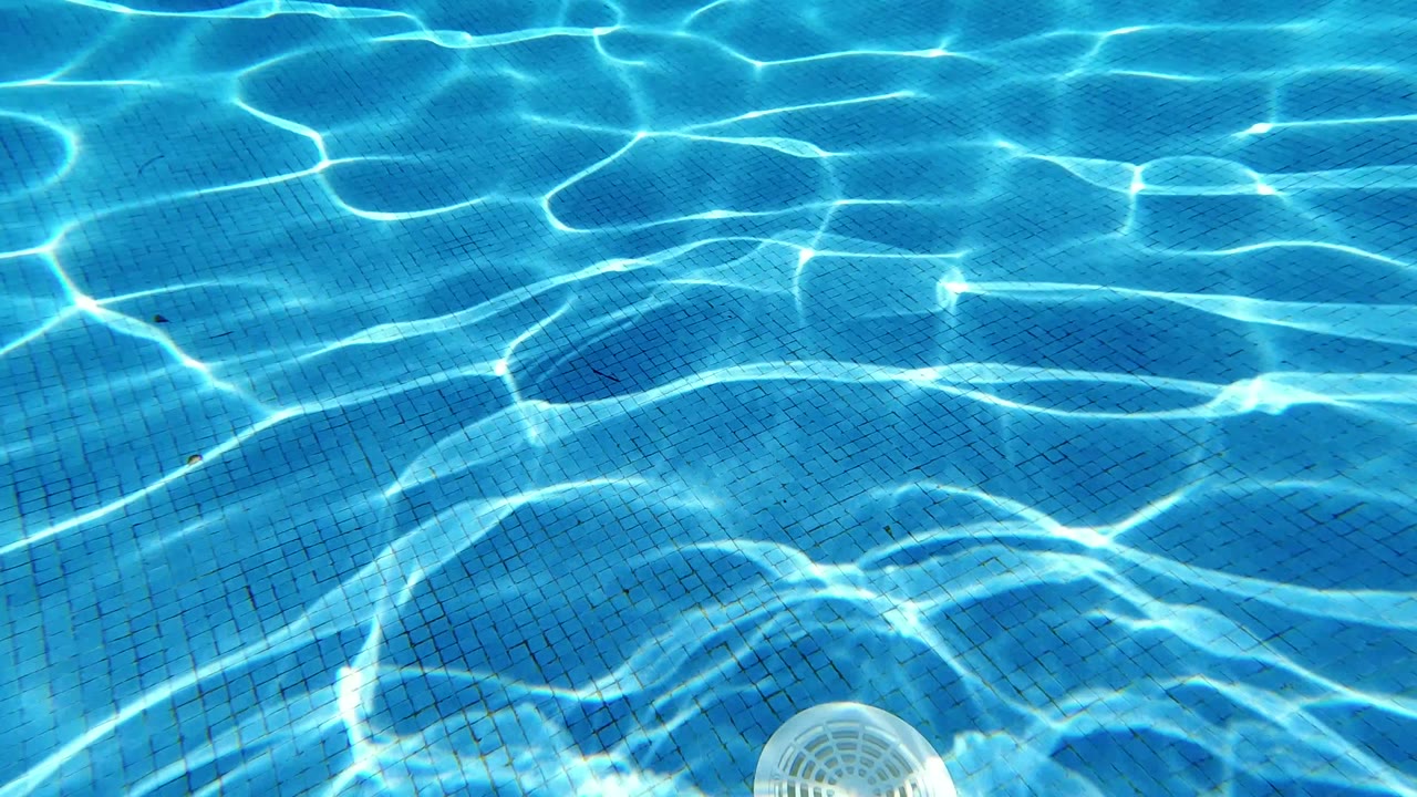 Clear blue pool water in Los Cabos with bright sun ripples over tile, a white drain near the lower left, and no visible people.