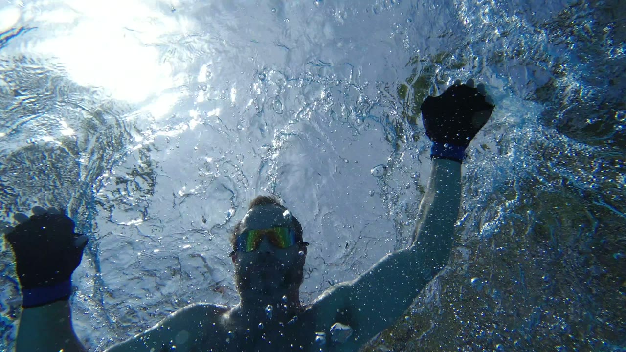 The swimmer has moved out of frame, leaving a Los Cabos underwater view of bright sun glare, rippled surface texture, bubbles, and blurred greenery above the water.