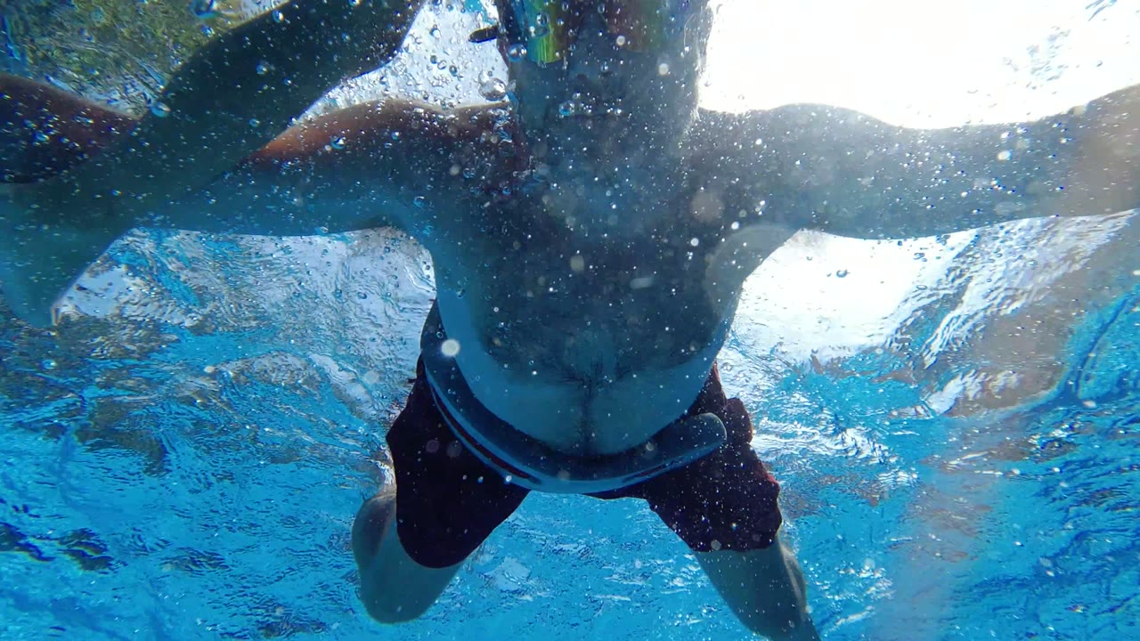 Underwater pool frame in Los Cabos showing a swimmer in dark trunks close above the camera, with blue water, bubbles, and bright surface light.