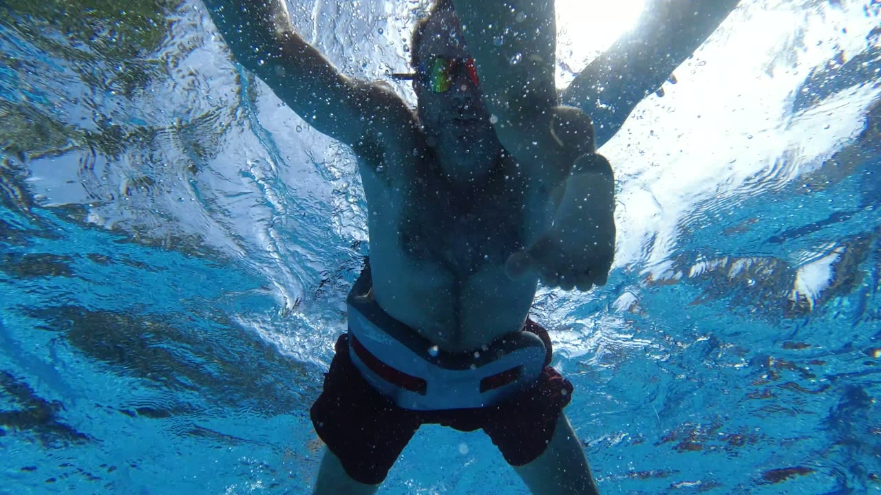 A swimmer passes directly above the underwater camera in Los Cabos, with arms extended, dark trunks, bubbles, and rippled sunlight across the pool surface.