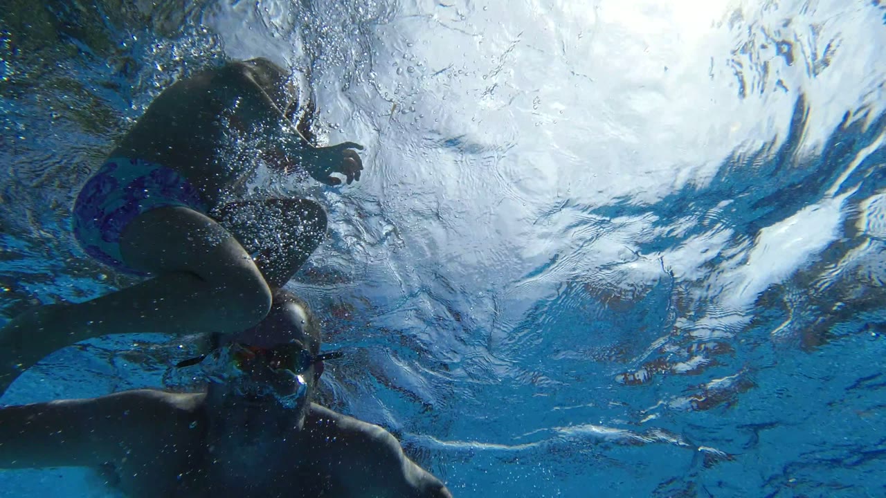 Underwater Los Cabos frame with an adult and child partly silhouetted near the surface, bubbles at left, and bright rippled water overhead.
