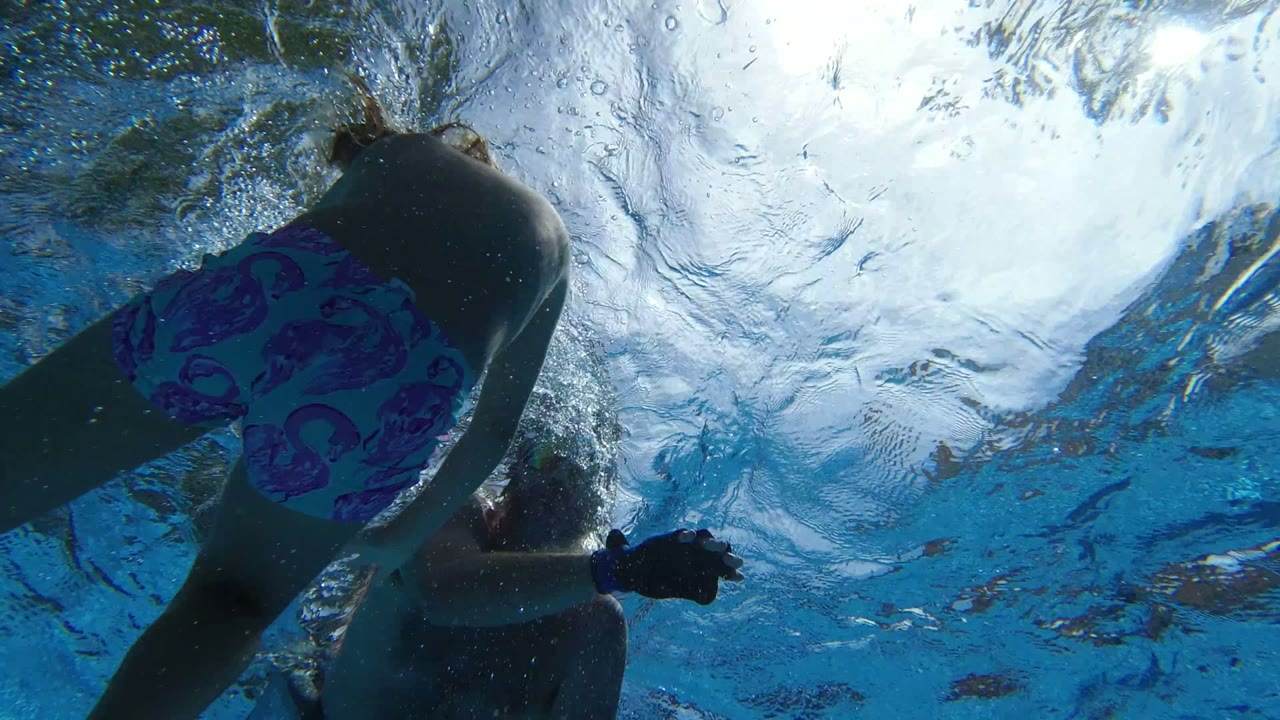 A child in a pink and blue swimsuit moves across the center of an underwater Los Cabos shot, with an adult partly visible nearby and bright surface reflections above.