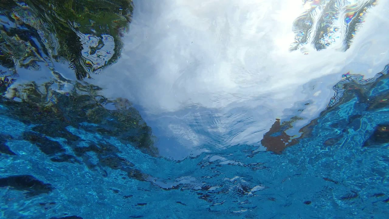 White surf and blue water shift across dark submerged shapes in Los Cabos, Mexico, with no visible people.