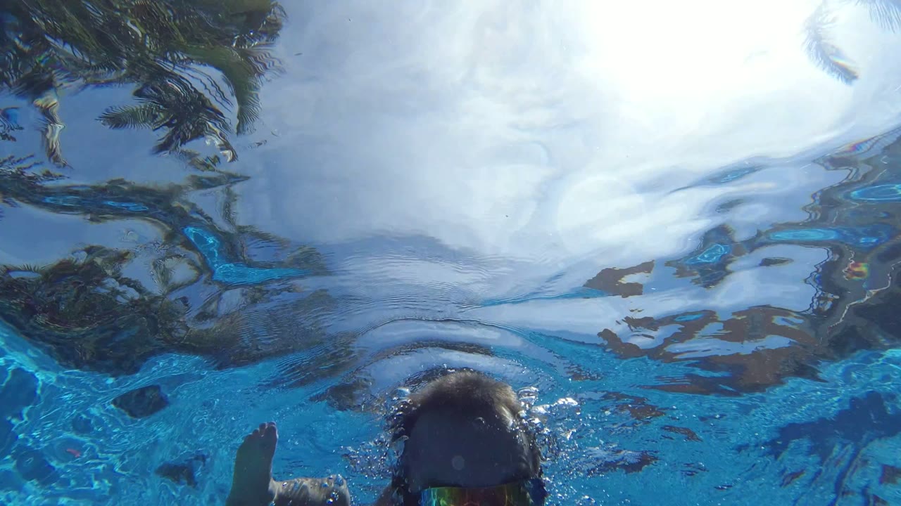 Underwater view in Los Cabos, Mexico, with a man in sunglasses centered near the surface, a partial child at left, and a swimmer in a flotation vest at right.