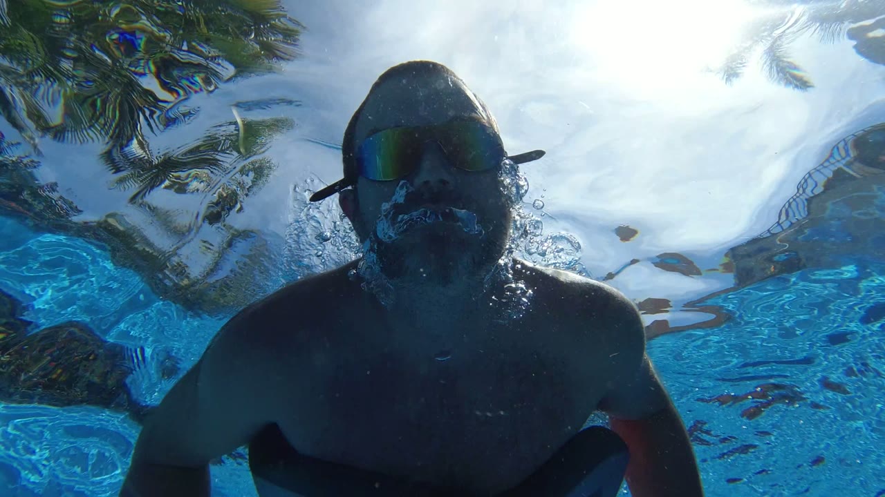 A swimmer wearing a flotation vest and blue hand paddles passes close over the underwater camera in Los Cabos, Mexico, with rippled surface light behind them.