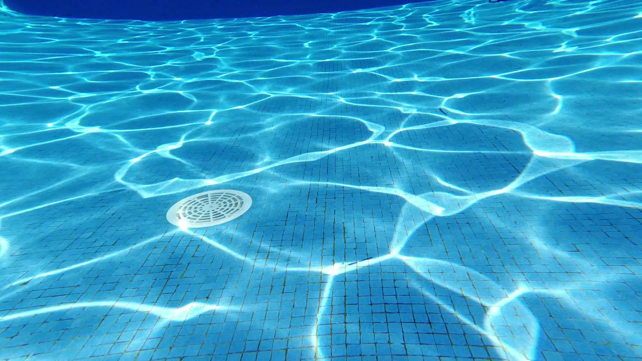 One second into the Los Cabos footage, sunlight ripples across the blue pool floor around a central drain cover, with no visible people.