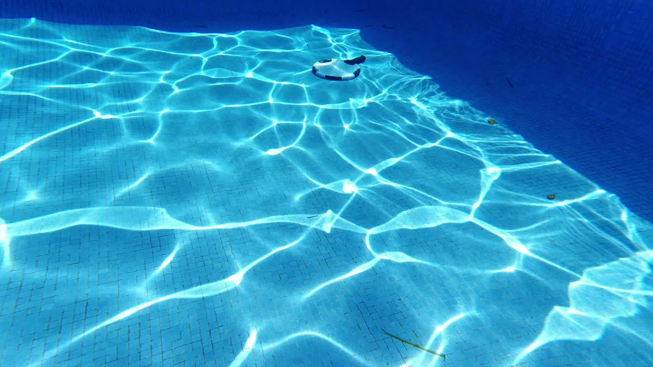 Overhead Los Cabos pool view with stronger sun patterns across the tiled bottom and the floating paddle set near the upper center, no visible people.