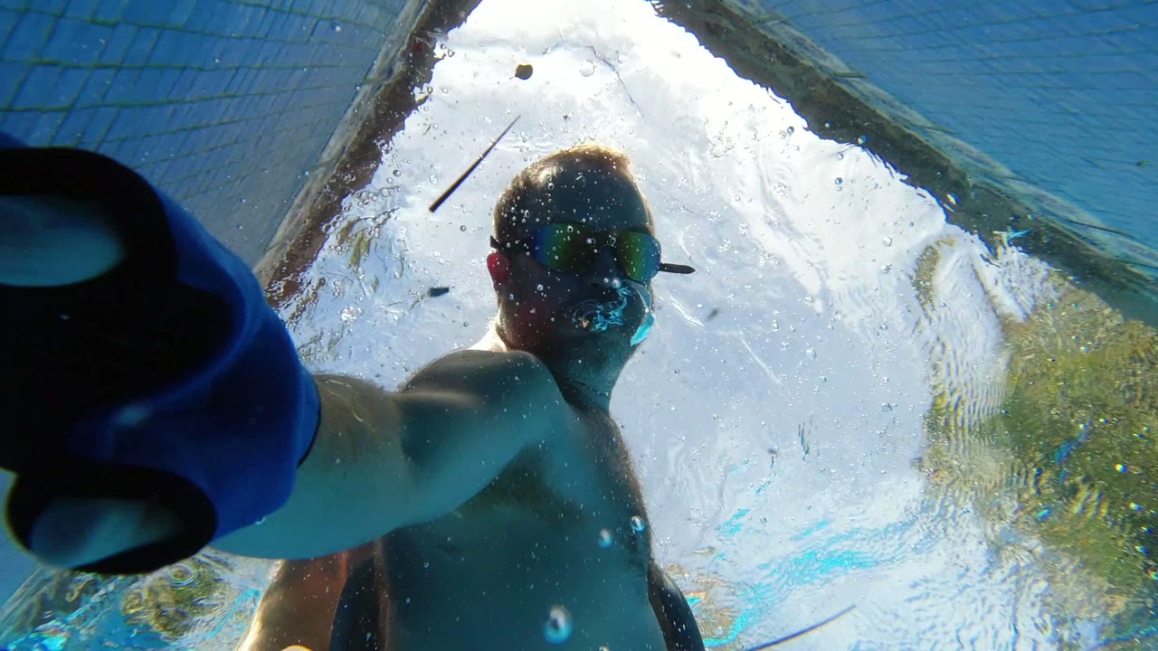 At 0:01 in Los Cabos, Mexico, a shirtless man wearing goggles reaches down toward the submerged camera through blue pool water and rippled sunlight.