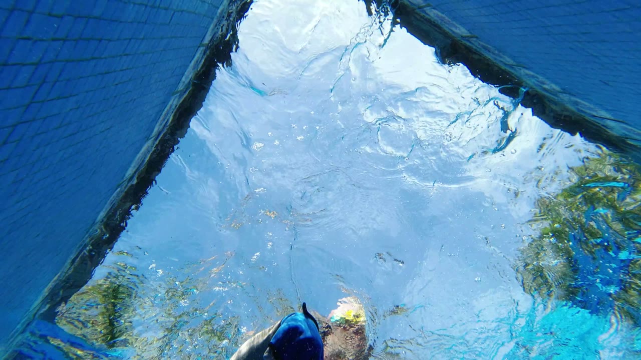 A partial swimmer remains near the bottom of the Los Cabos underwater frame, with bubbles, blue water, sky reflection, and a dark diagonal pool edge above.