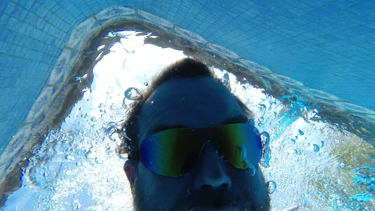 A goggled adult man is framed underwater in Los Cabos, with sunlight breaking through the rippled pool surface and bubbles around his face.