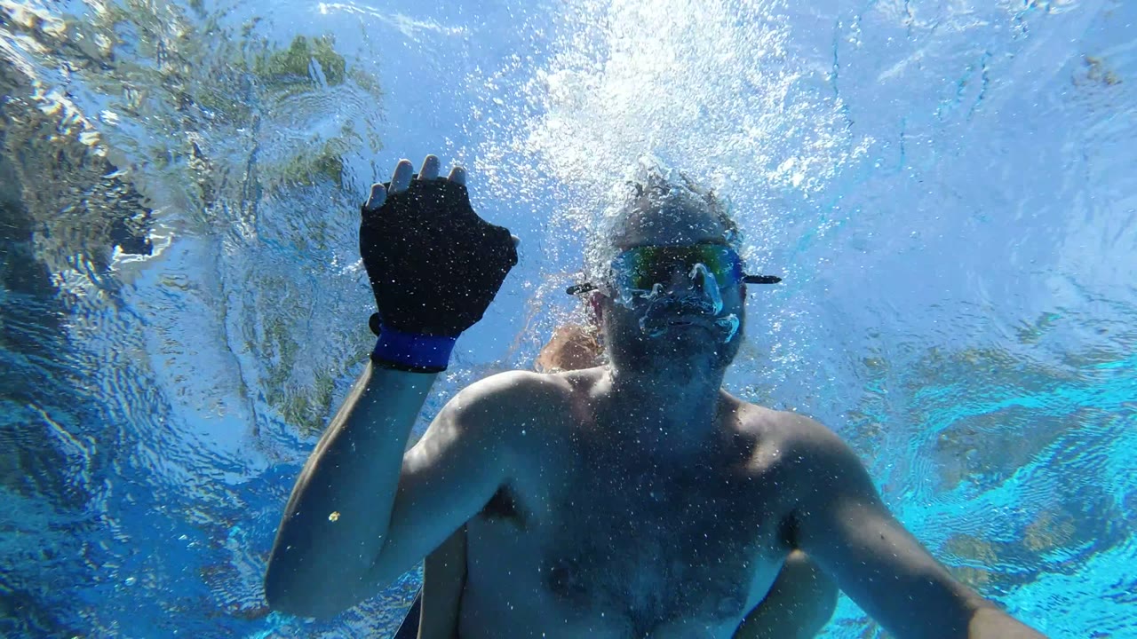 The adult swimmer in goggles raises a blue-gloved hand below the water surface in Los Cabos, Mexico, with bubbles and reflected light around his face.