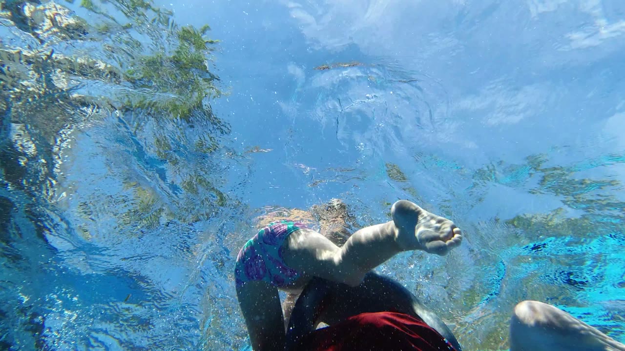 Underwater view in Los Cabos, Mexico, looking up through blue pool water as bare legs and red fabric appear near the lower edge.