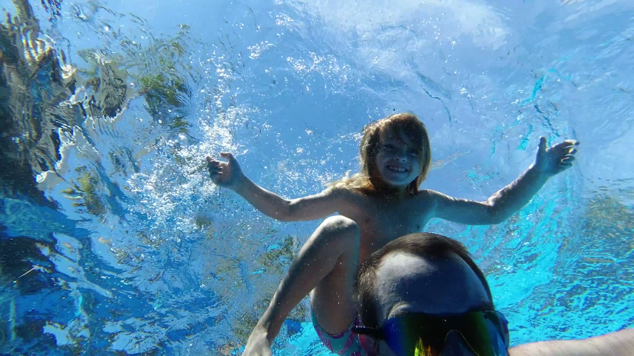 Child above an adult man underwater in a blue swimming pool.