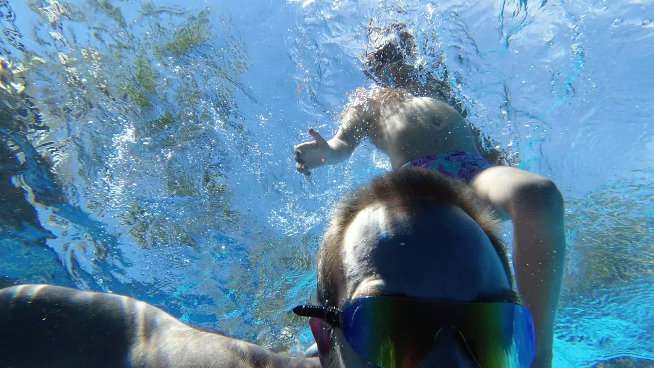 In Los Cabos, the adult man is close to the submerged camera while another swimmer passes above him through rippled pool water.