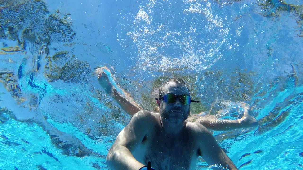 At 0:02, the adult swimmer remains underwater in the Los Cabos pool, shifted toward the right side with rippled surface reflections and bubbles above.