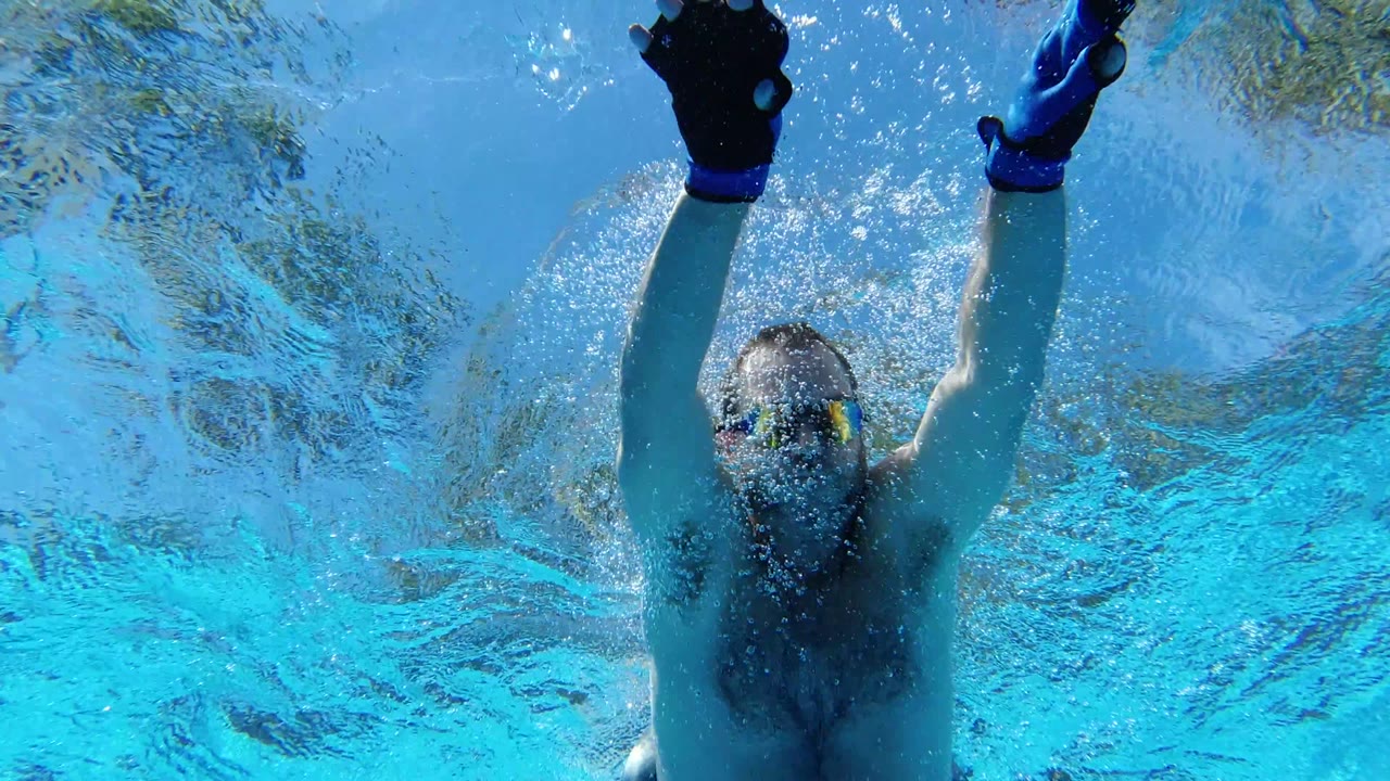 At 0:00 in Los Cabos, Mexico, an adult man wearing swim goggles is underwater with both arms raised near the rippled pool surface.