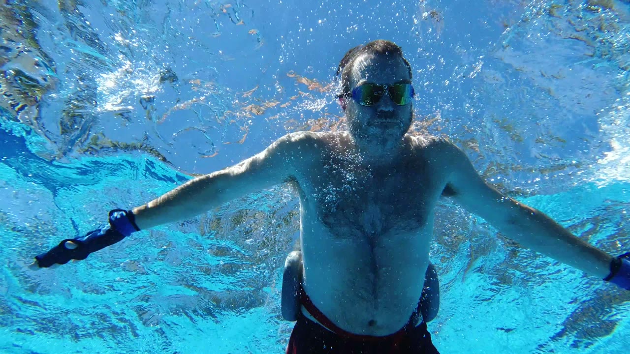 At 0:01 in Los Cabos, Mexico, the goggled swimmer is centered underwater with arms extended sideways beneath the shimmering pool surface.