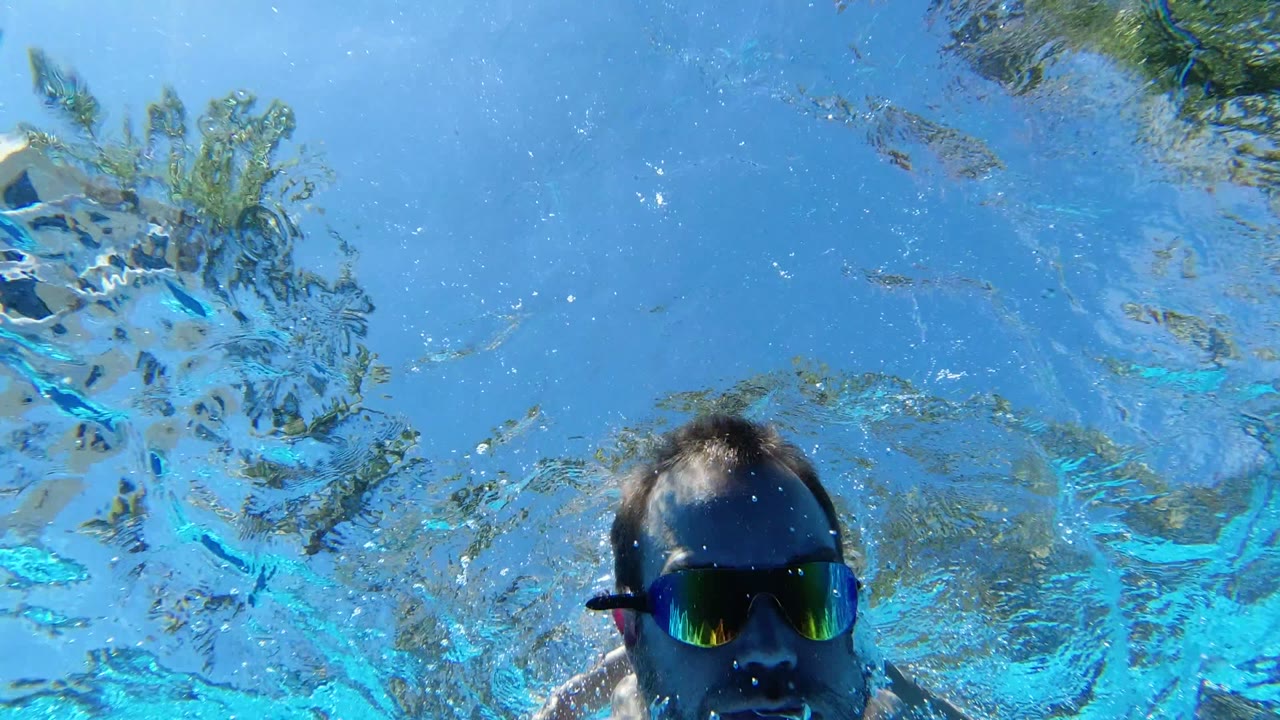 A shirtless adult man in mirrored swim goggles moves toward the underwater camera in a Los Cabos pool, with distorted reflections and blue water around him.
