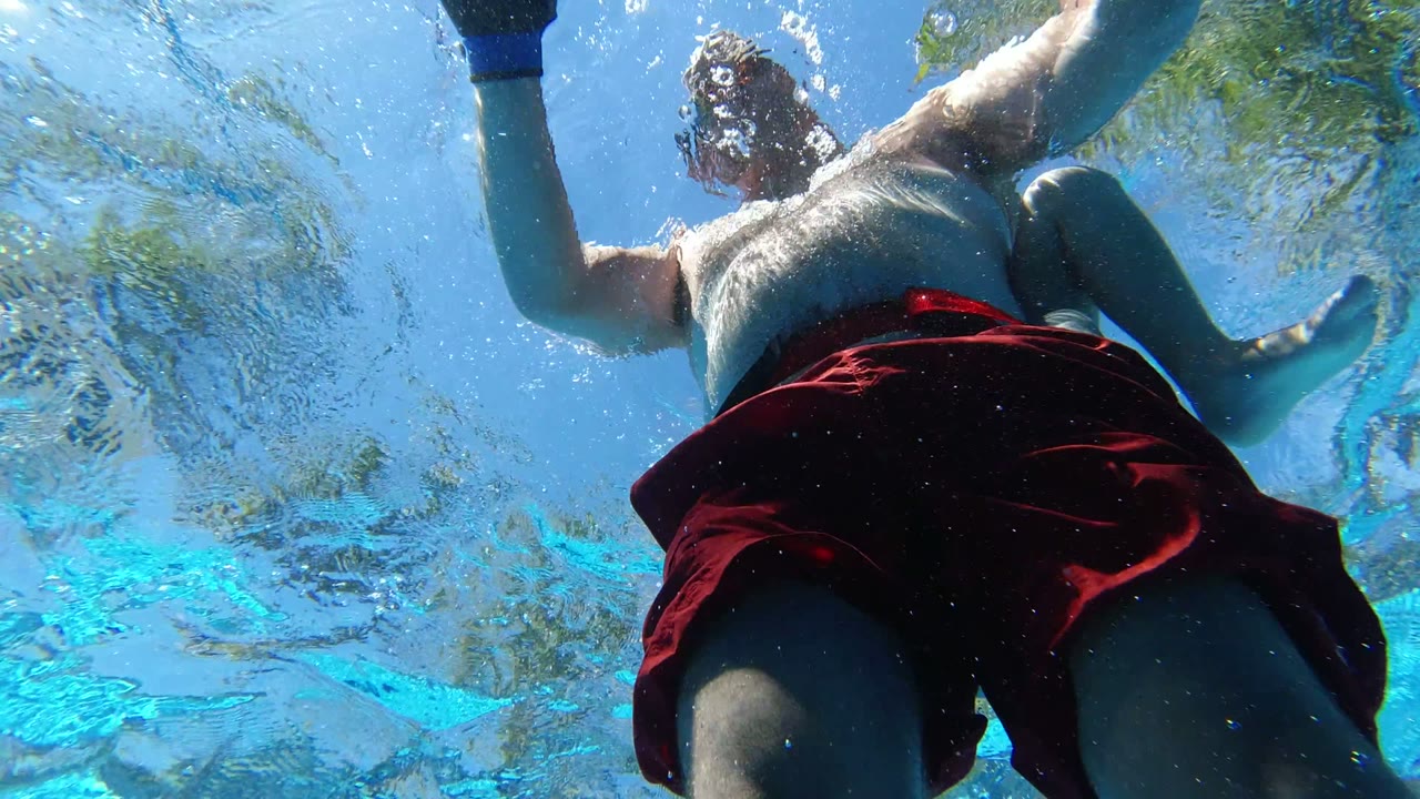 Underwater Los Cabos pool view of the swimmer centered overhead, with red trunks, pale torso, raised arms, bubbles, and sunlit surface ripples.