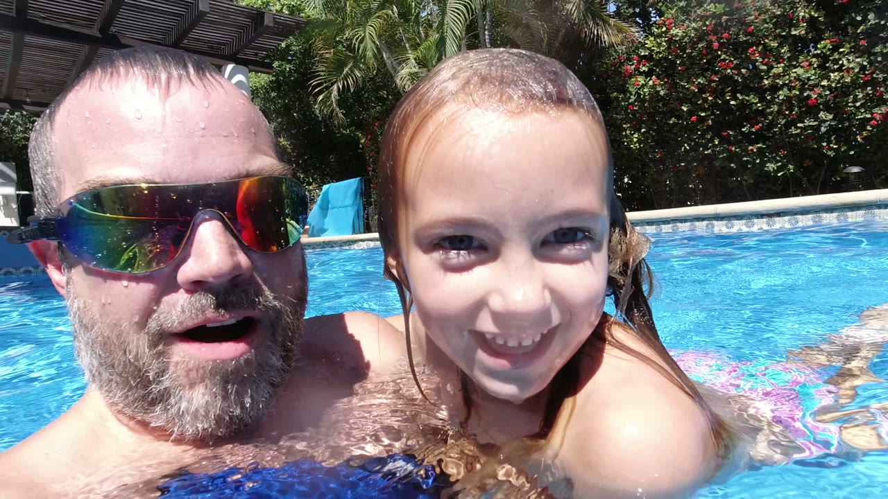 The man and young girl smile near the waterline in the Los Cabos pool, with bright sunlight on their wet faces and a shaded structure in the background.
