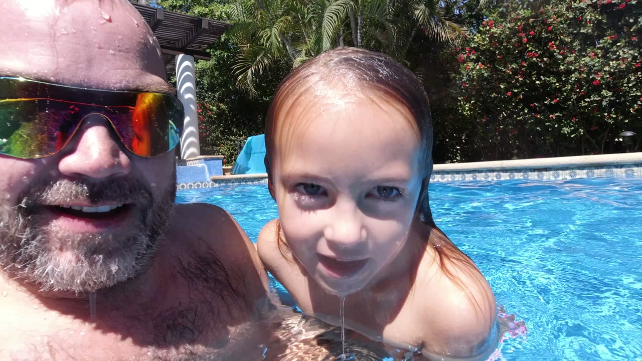 The father smiles beside the young girl at water level in the Los Cabos pool, with rippled blue water, tiled pool edge, greenery, and shade columns behind them.