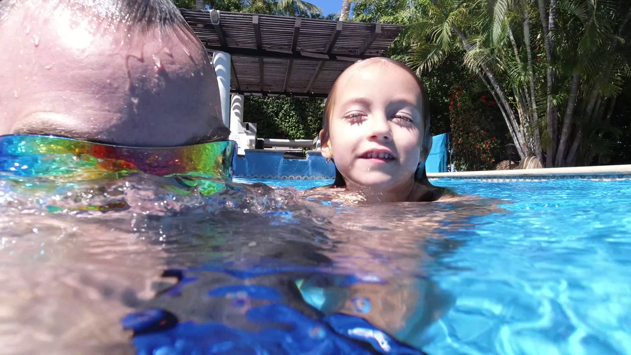 At the waterline in Los Cabos, Mexico, the man's mirrored sunglasses sit half above the rippling surface while the child floats nearby in bright blue pool water.