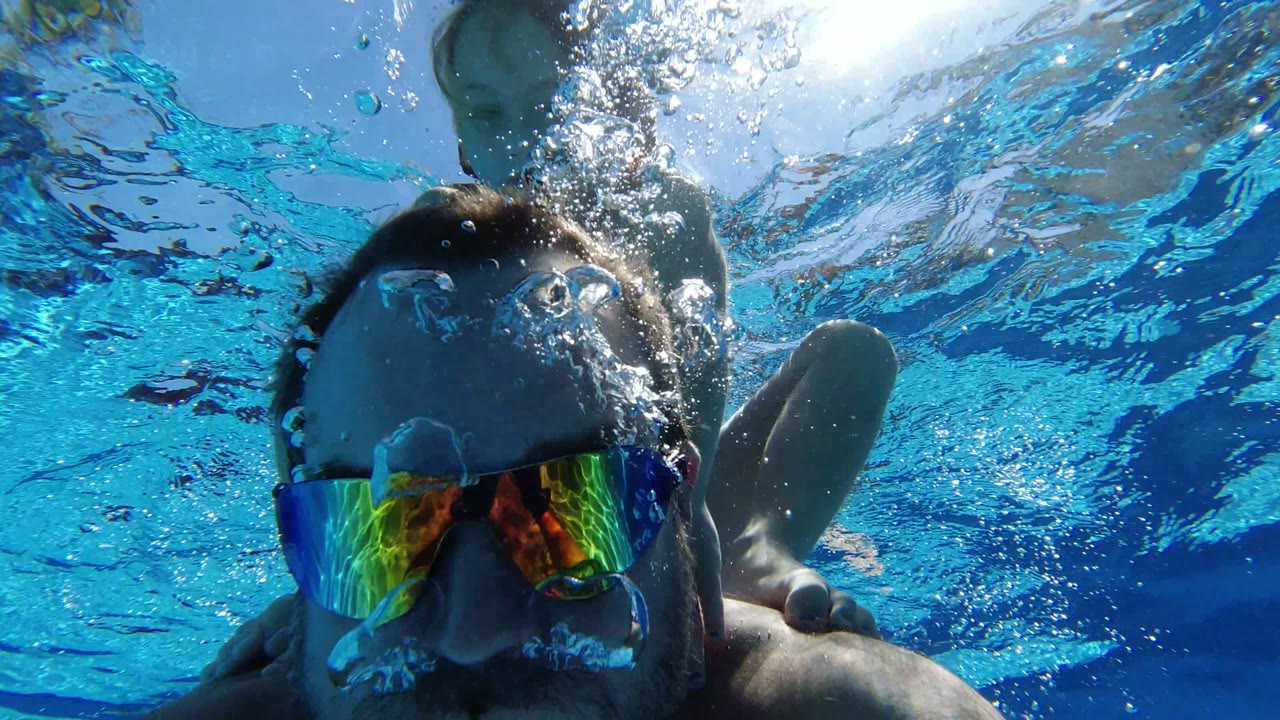 At 0:01 in Los Cabos, Mexico, the man stays close to the camera while a child is partly visible behind his shoulder and bubbles break across the water surface.
