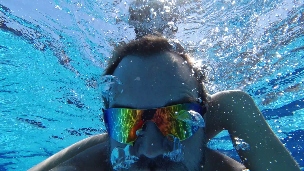 Close underwater view in Los Cabos, Mexico of a bearded adult swimmer wearing mirrored goggles, with blue pool water and surface ripples above him.