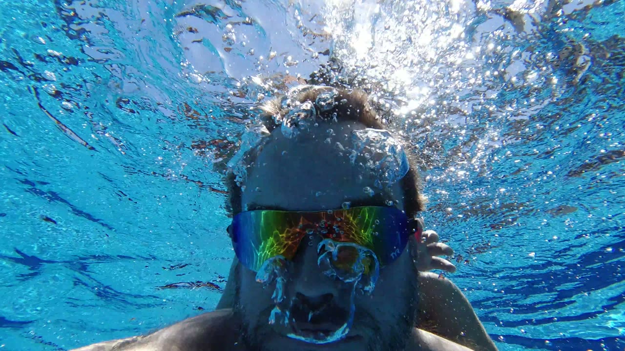 The swimmer remains centered underwater in Los Cabos, Mexico, with bubbles crossing his forehead and reflected color in the mirrored goggles.