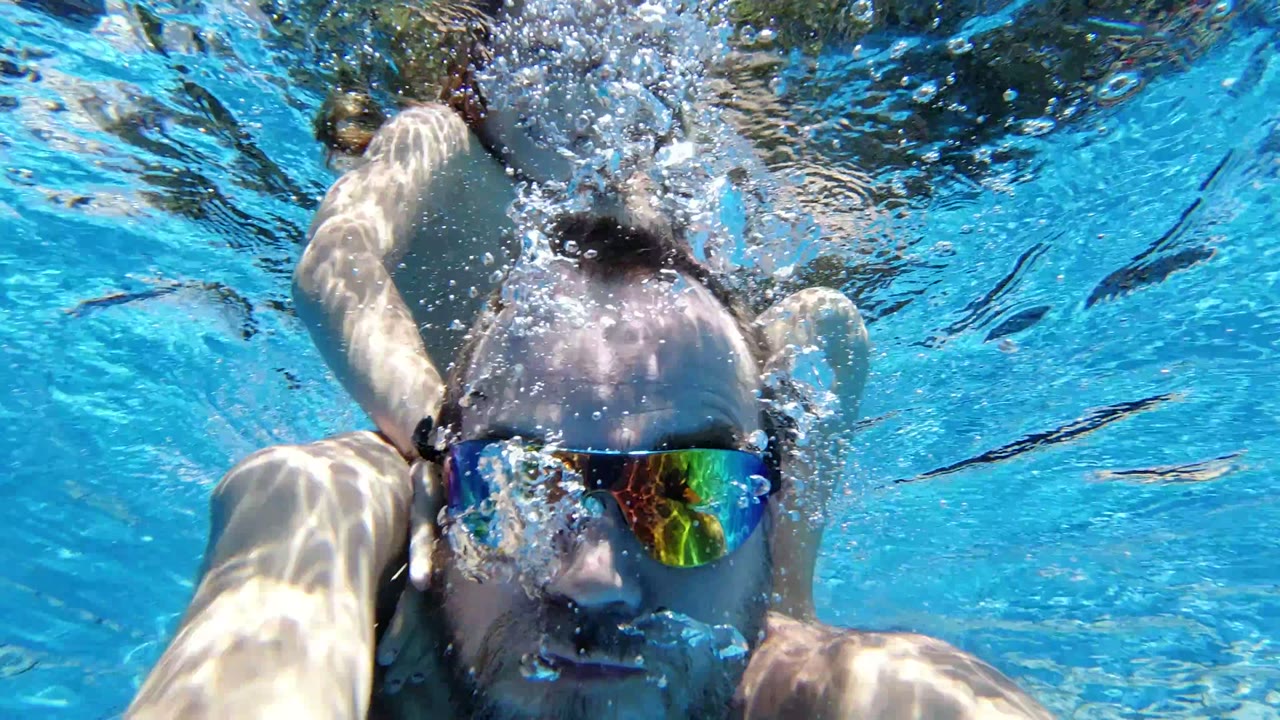 Adult man underwater in mirrored goggles with bubbles in a blue pool.