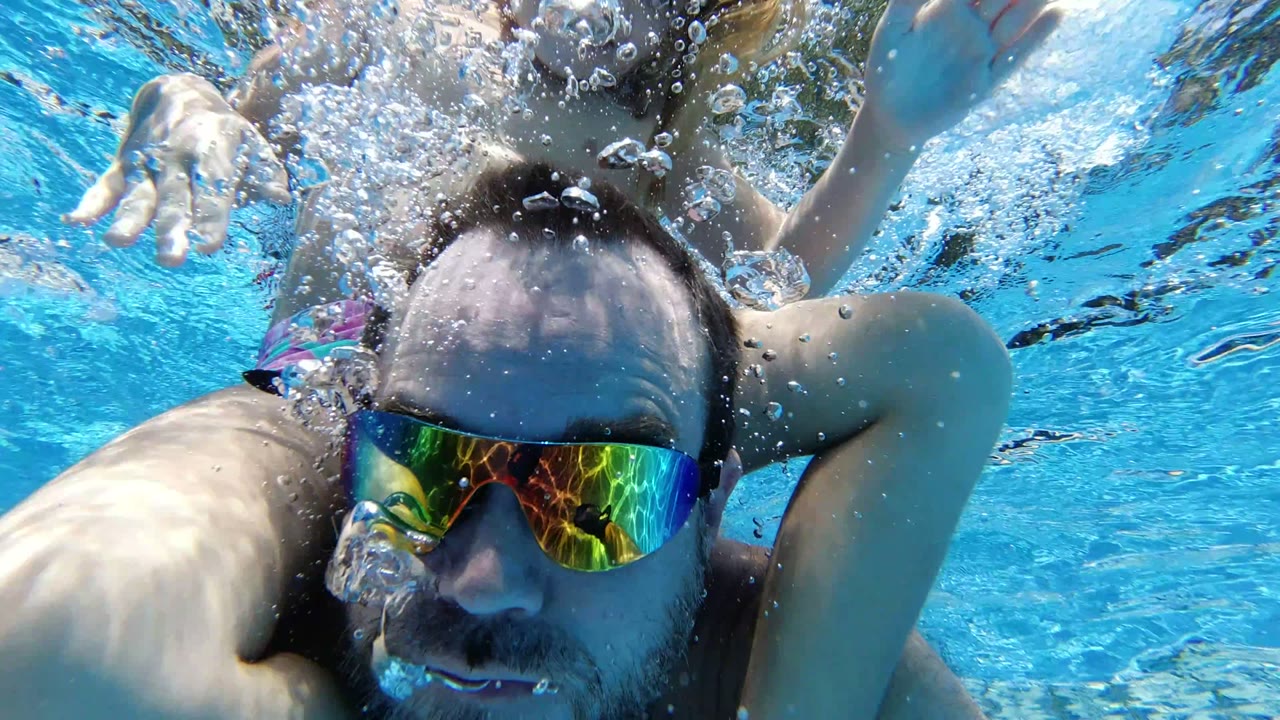 At 0:01 in Los Cabos, Mexico, the adult man's goggled face stays close to camera while a child's arm and swimsuit appear behind him in the blue pool water.