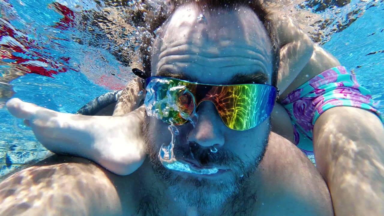 The father's goggled face stays close to the underwater camera as the child's foot and legs pass across the upper frame in the Los Cabos pool.