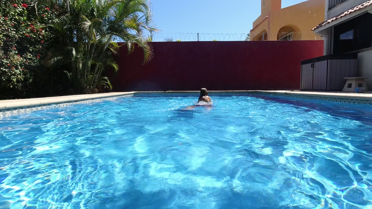 The swimmer moves across the far side of the Los Cabos pool while sunlight creates bright ripples across the water and the red wall stays in the background.