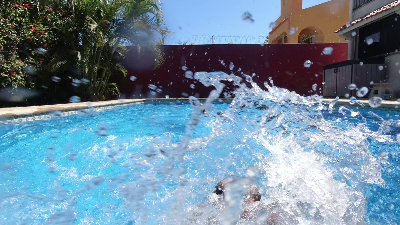 A large burst of pool water fills the foreground in Los Cabos, Mexico, with blue water, a red backyard wall, palm trees, and partial swimmers obscured by the splash.