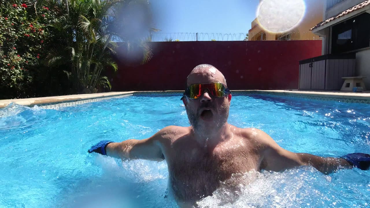 An adult man wearing mirrored swim goggles surfaces close to the water-level camera in a bright blue pool in Los Cabos, Mexico, with water splashing around his chest.