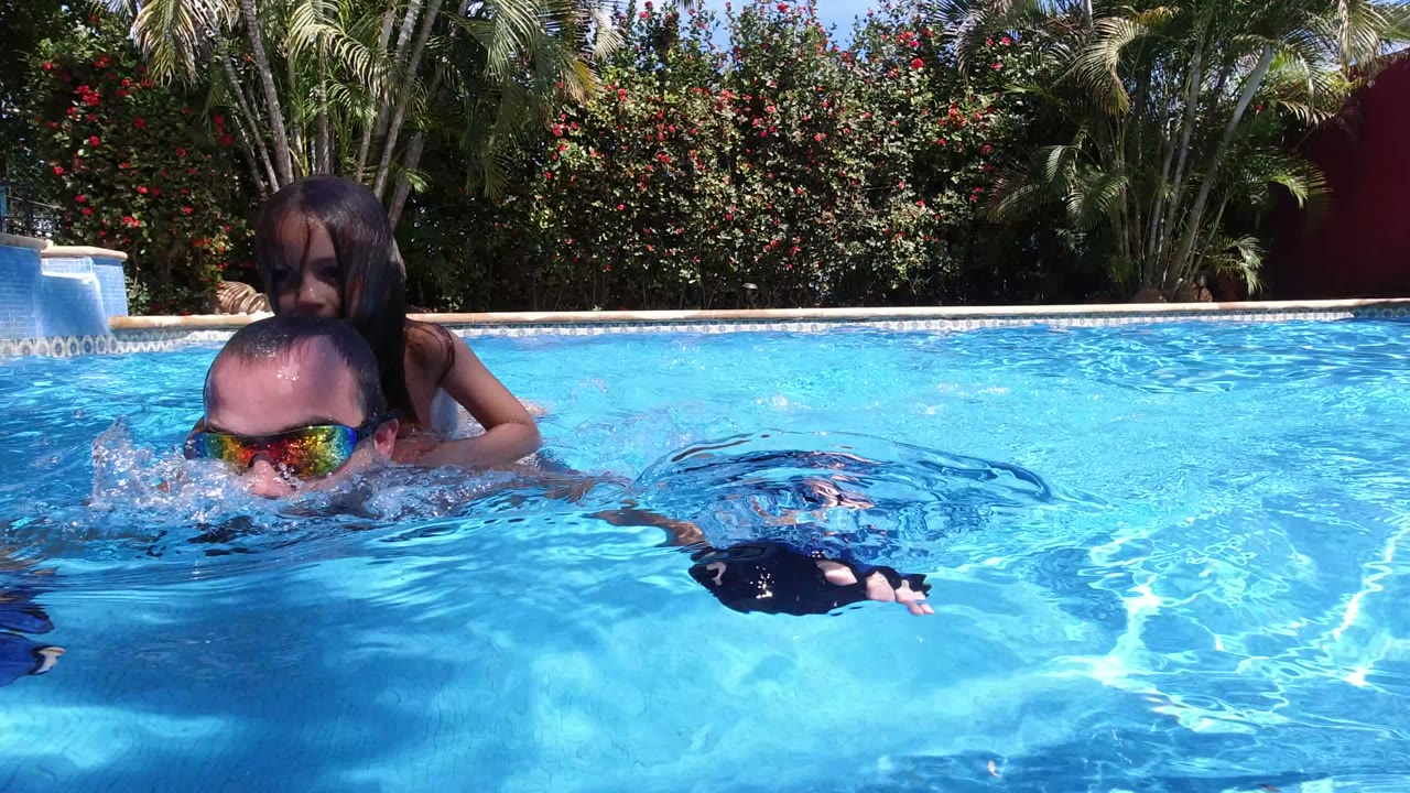 A young girl sits behind an adult man in reflective swim goggles as they move through a Los Cabos swimming pool under bright sunlight.