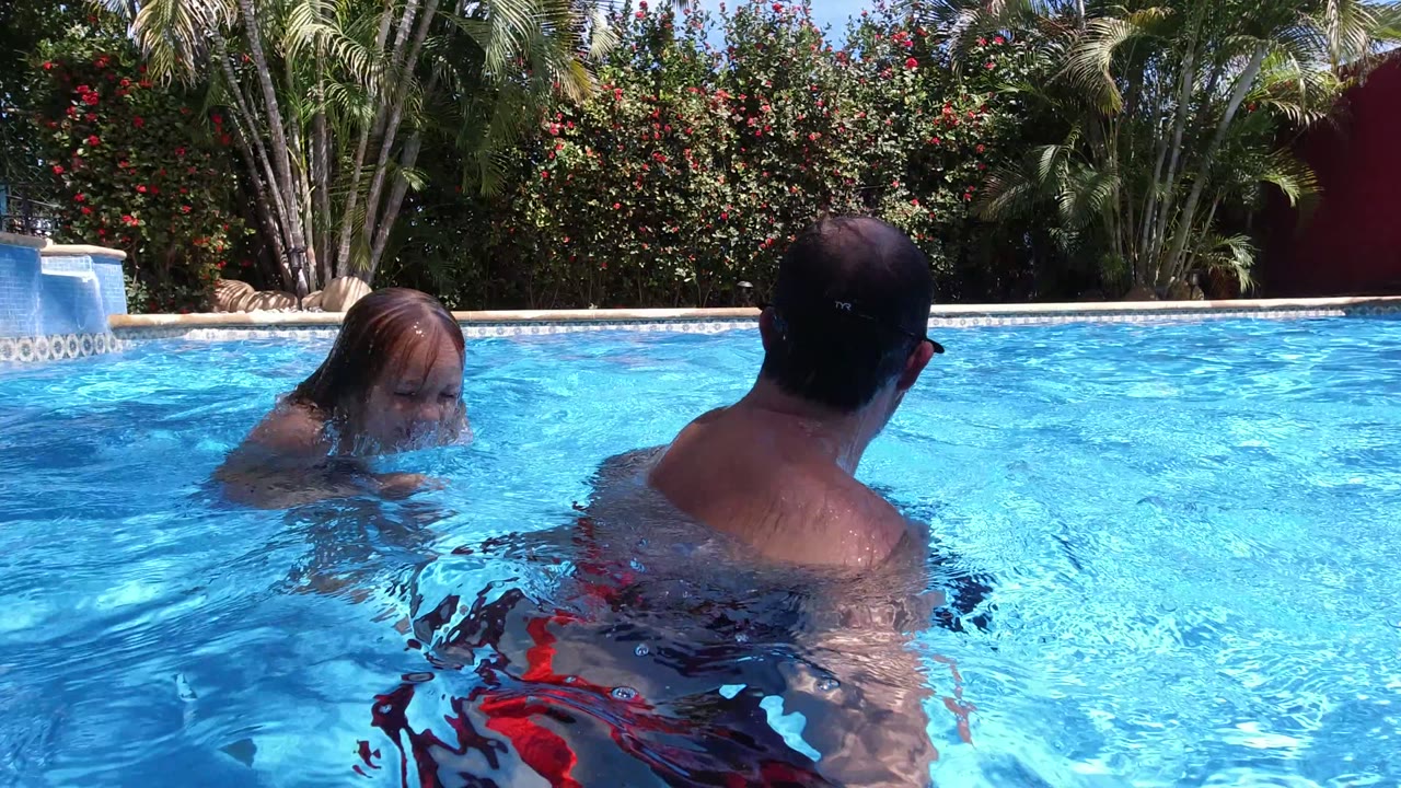 A young child faces a man from across the bright blue pool water in Los Cabos, with green trees and a low pool edge in the background.