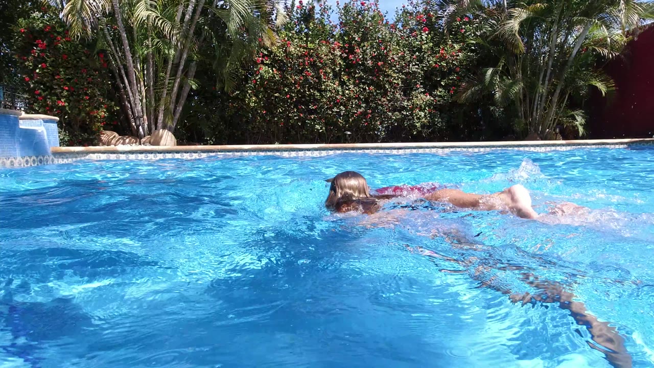 A swimmer surfaces while moving across the blue pool in Los Cabos, Mexico, with ripples, splashes, palms, and a hedge behind them.