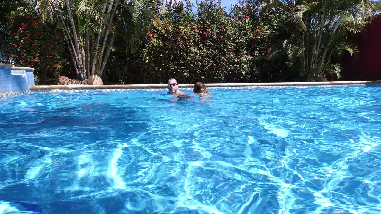 In Los Cabos, Mexico, a man and child are partly above the surface of a bright blue backyard pool, with palms and flowering hedges behind the far edge.