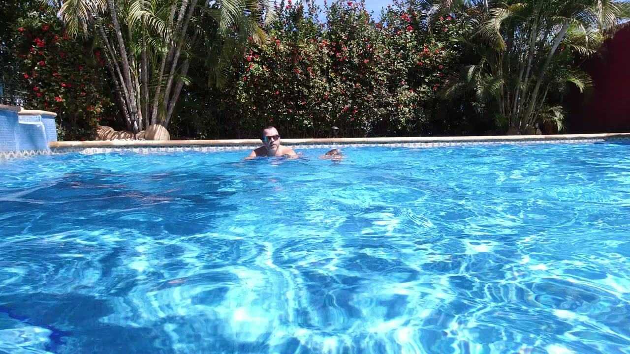 In Los Cabos, Mexico, the man faces toward the camera from the pool while the child is lower in the water to his right, with bright reflections across the foreground.
