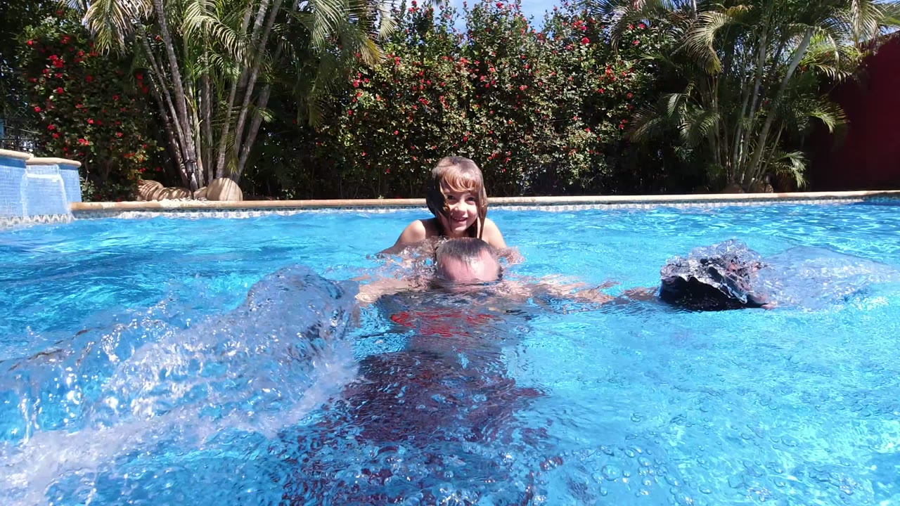 In Los Cabos, Mexico, a child smiles behind an adult partly submerged in a bright blue pool as water splashes toward the camera and palms fill the background.