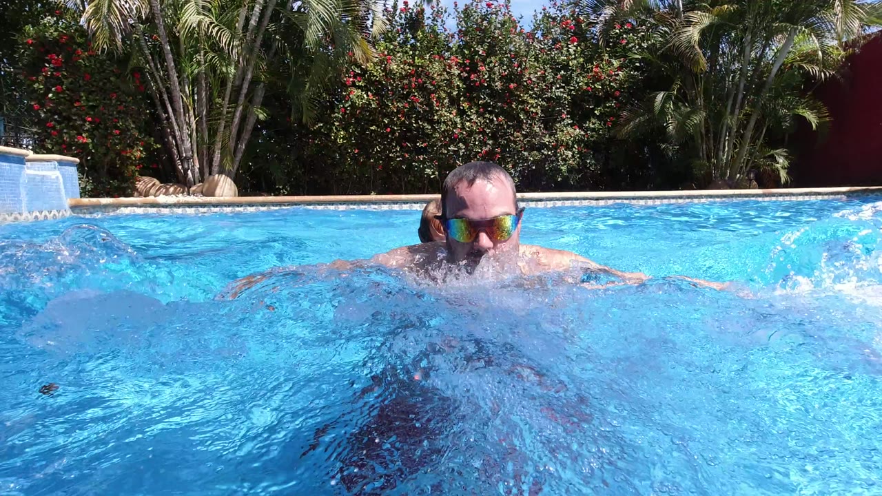 In Los Cabos, Mexico, an adult man wearing reflective sunglasses surfaces at water level while splashes partially obscure his torso and the landscaped pool edge sits behind him.