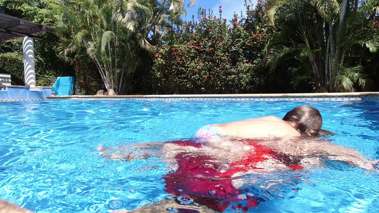 At the start of the Los Cabos clip, a child lies across a man's back as he swims close to the water-level camera in a bright blue pool.