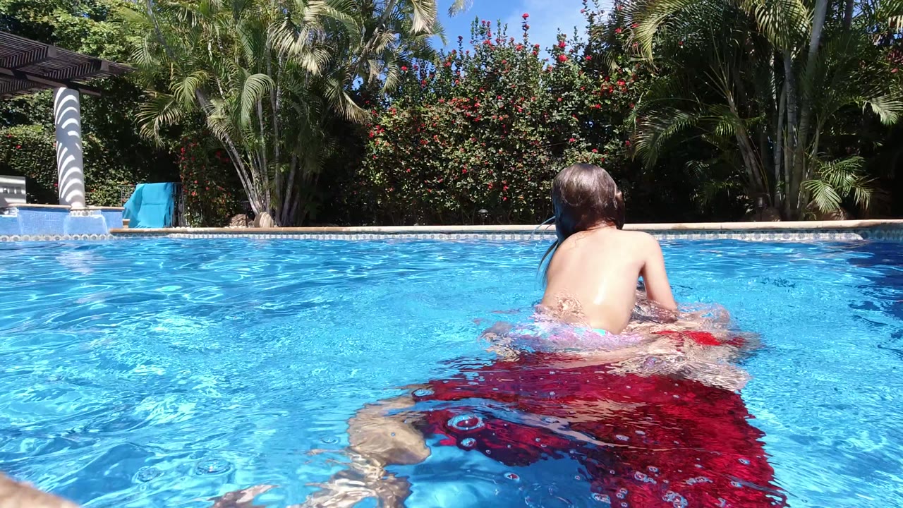 One second in, the man and child have moved farther from the camera in the Los Cabos pool, with palms and red-flowering shrubs behind them.