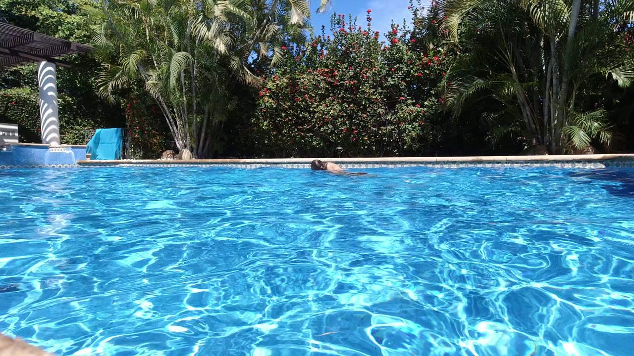 One second in, the swimmer is farther across the Los Cabos pool, with rippled blue water dominating the frame and a white column and dense garden behind the deck.