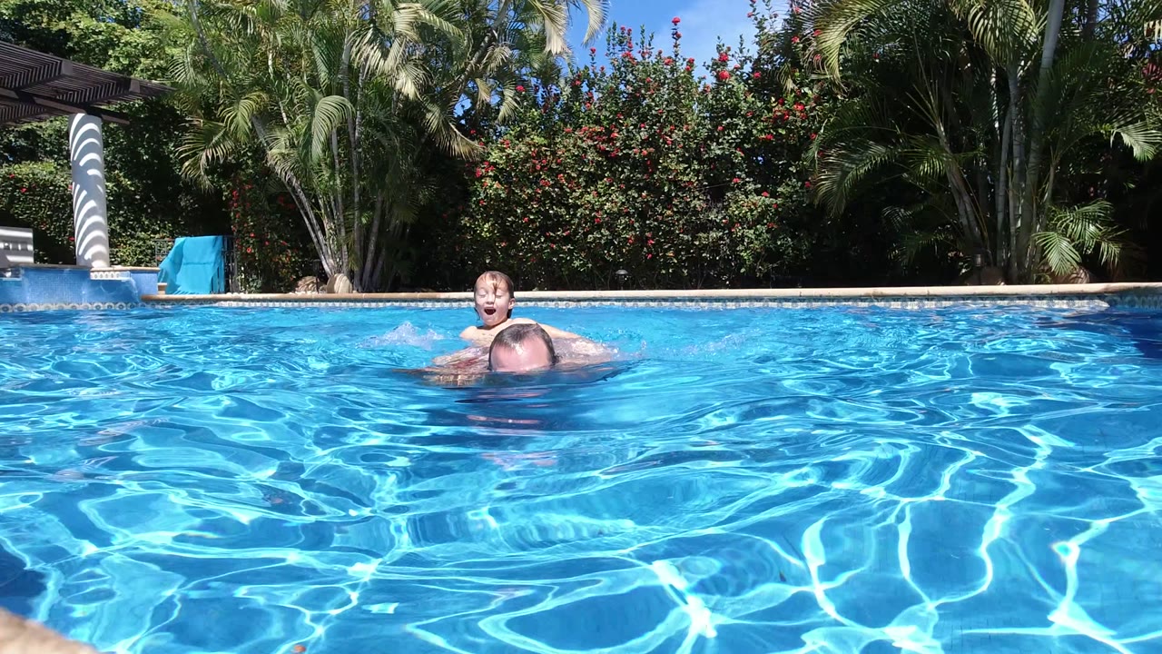 The young girl appears higher above the water behind the father as he swims forward in the Los Cabos pool, with sunlit water and dense greenery behind them.
