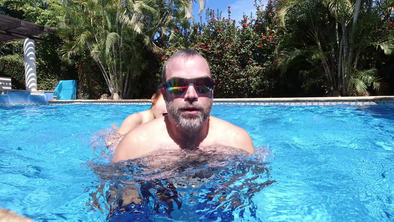 An adult man in sunglasses faces the camera at water level in a Los Cabos pool, with a child partly hidden behind him and palms and shrubs beyond the pool edge.